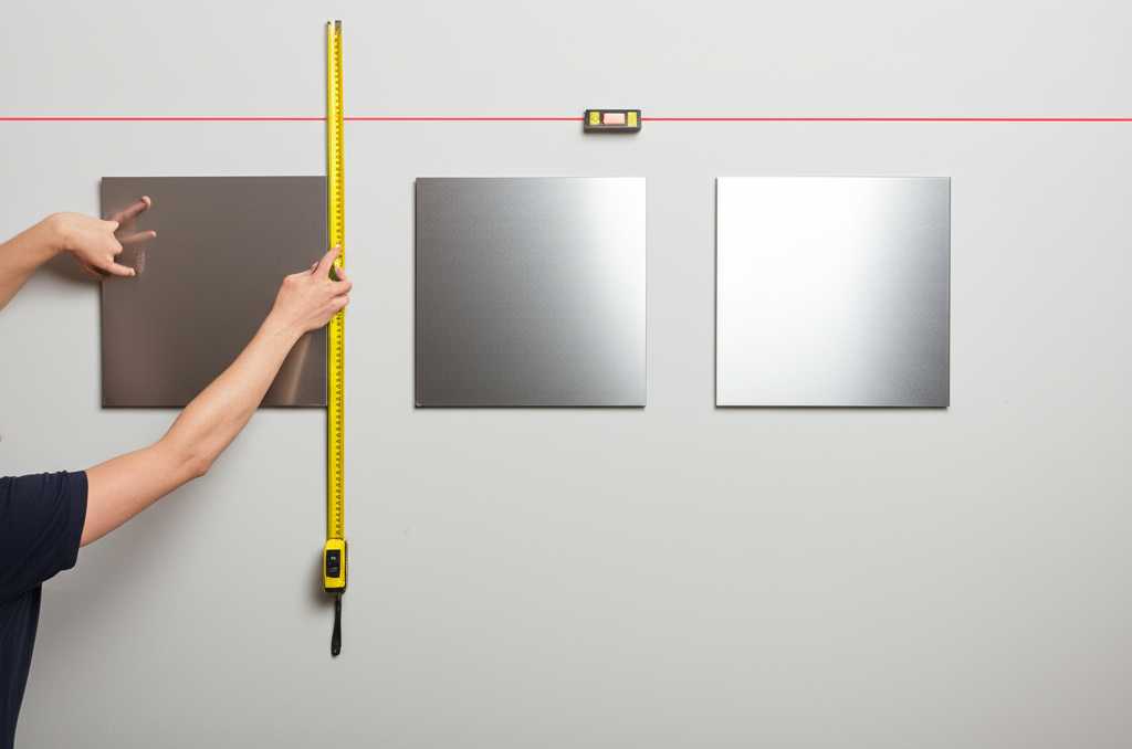 A person using a laser level and measuring tape to perfectly align three medium-sized metal prints on a clean wall, focusing on the act of precise installation.