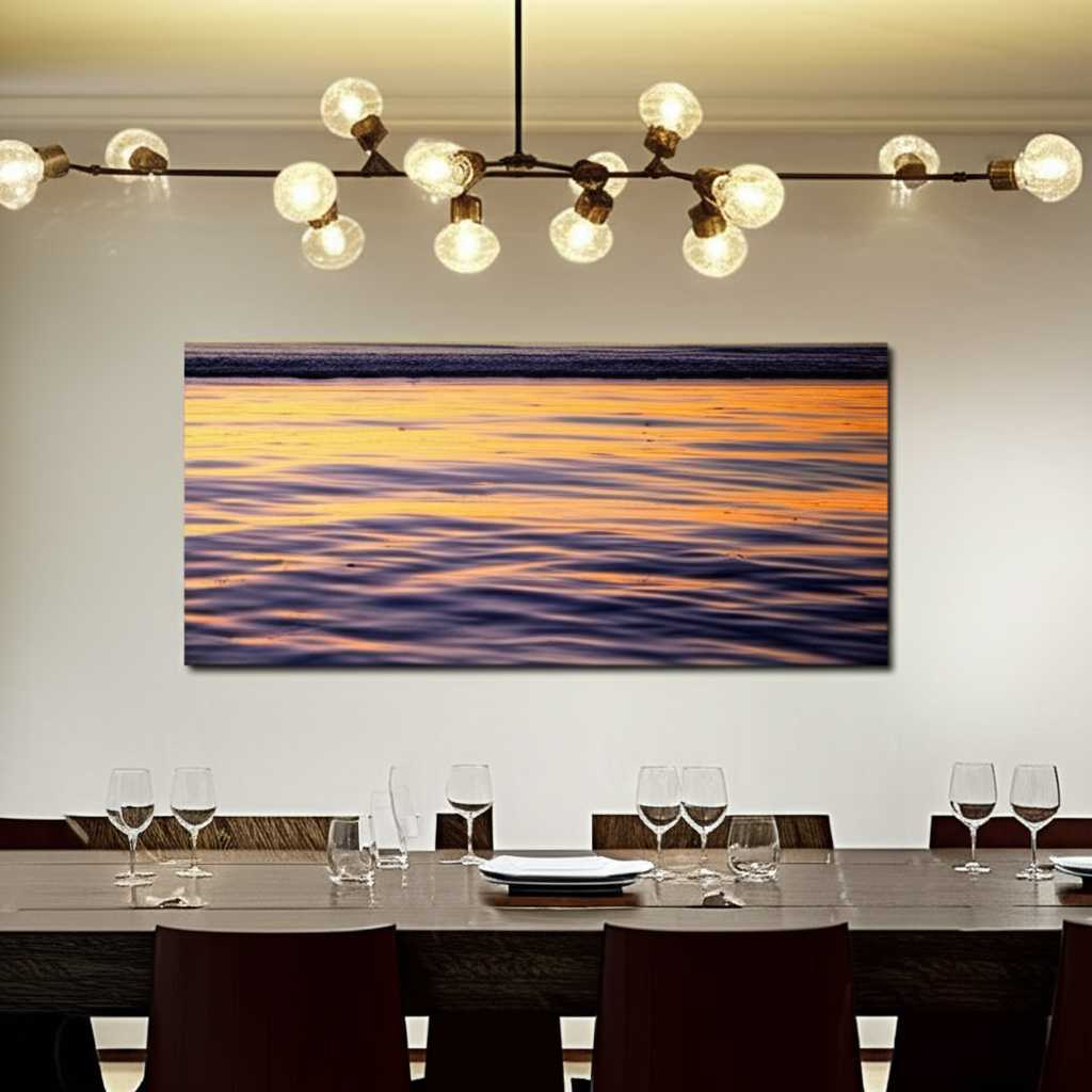 A panoramic metal print of a serene landscape hanging in a dining room, beautifully reflecting the warm light from a modern chandelier above the table.
