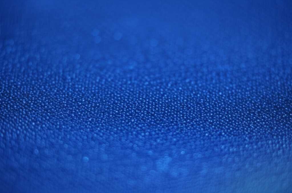 A close-up, angled shot of a minimalist metal art piece. The light is catching a subtle, UV-printed texture on the surface, creating an intricate play of light and shadow across a simple colored area.